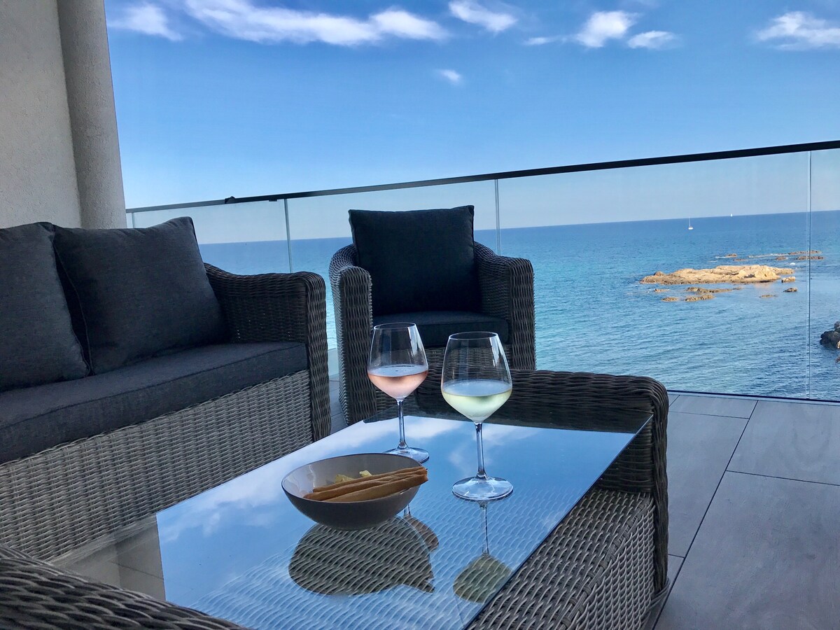 A balcony setup features two glasses of wine, one pink and one green, placed on a glass-topped table. Soft seating in light gray wicker surrounds the table, providing a view of the calm sea and blue sky in the background.