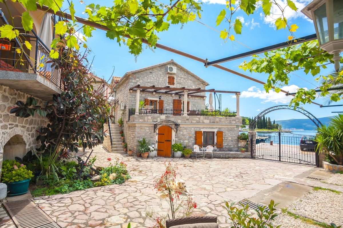 A traditional stone building is surrounded by greenery and colorful flora. A paved courtyard features various seating options. The entrance is framed by wooden windows and a cheerful orange door, while distant views of the Adriatic Sea can be glimpsed beyond the garden.