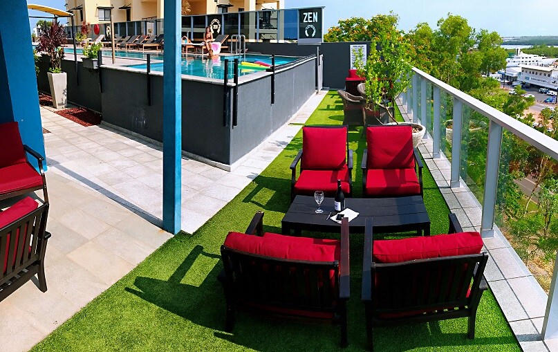 A rooftop lounge area features plush red seating with accompanying dark tables, set against vibrant green turf. The infinity pool is visible in the background, inviting relaxation while enjoying the surrounding views. Sunlight casts a warm glow over the entire space, enhancing the outdoor atmosphere.