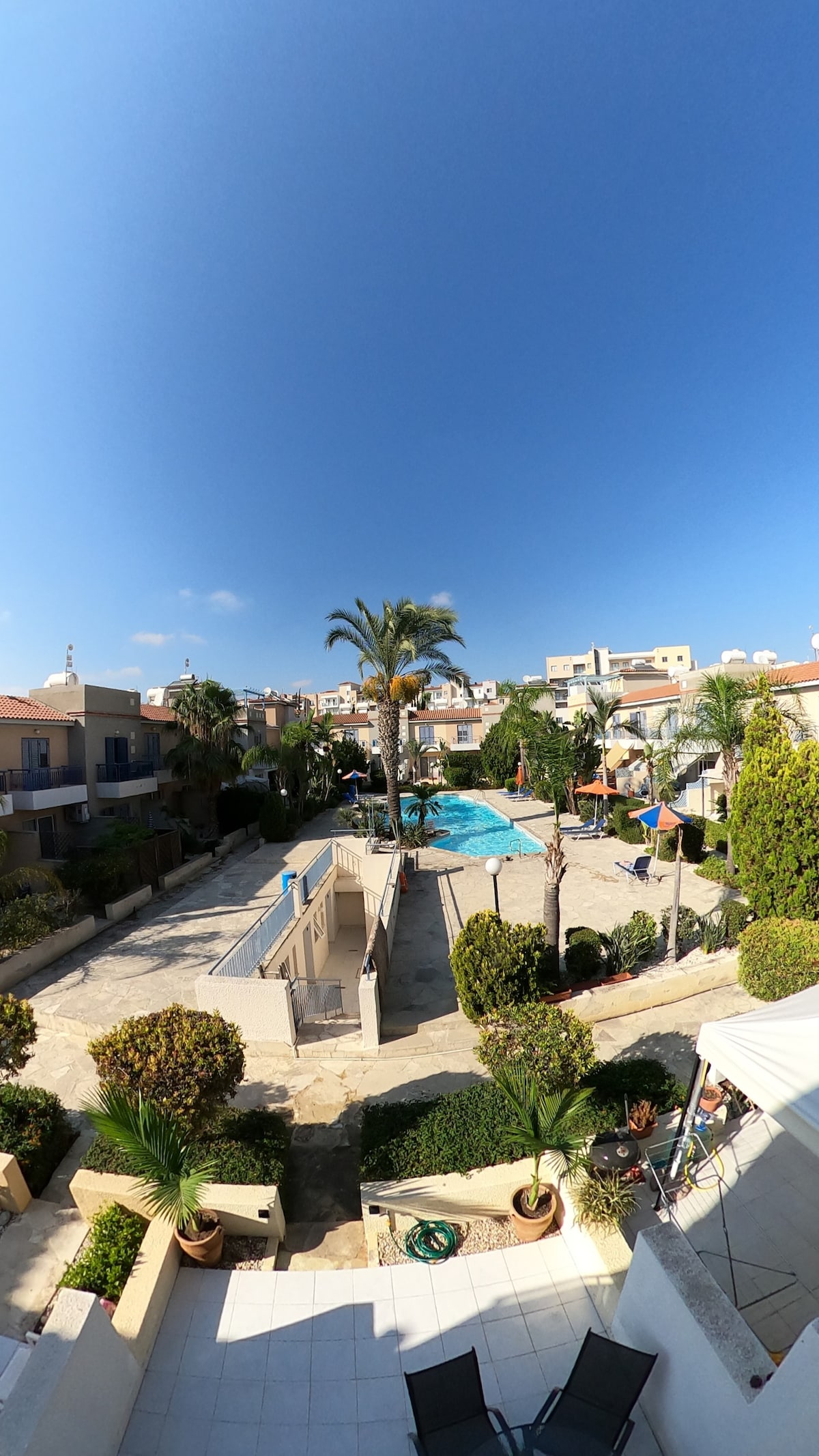 A wide view reveals a serene outdoor space featuring a swimming pool surrounded by palm trees and lounge areas. The manicured gardens and pathways lead to charming accommodations, with vibrant colors and clear skies enhancing the peaceful setting.