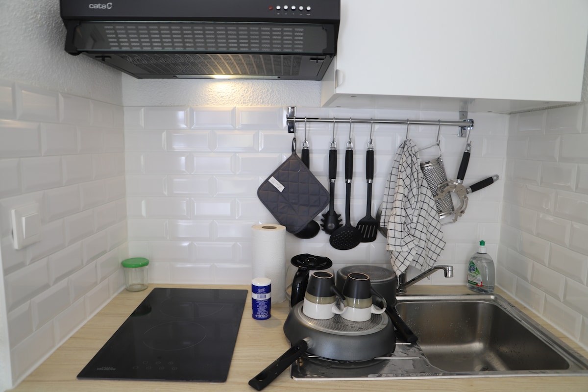 A functional kitchen area is showcased, featuring a sleek countertop with a built-in stovetop and a sink. Kitchen utensils hang on a rack above, complemented by a pot and various cleaning supplies nearby. White tiles enhance the bright and practical atmosphere.