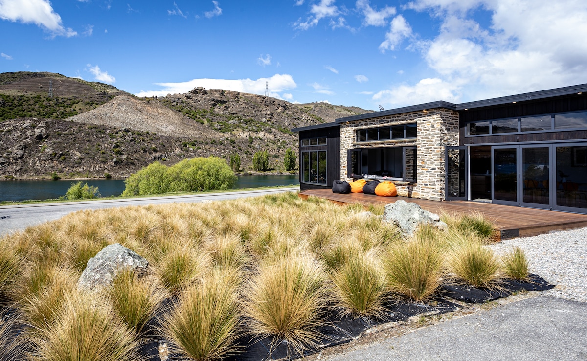 A modern house sits beside a lake, featuring a spacious deck and large windows that invite natural light. Surrounding the home are neatly arranged native grasses and boulders. The scenic backdrop includes rolling hills and a clear blue sky, enhancing the overall serene environment.