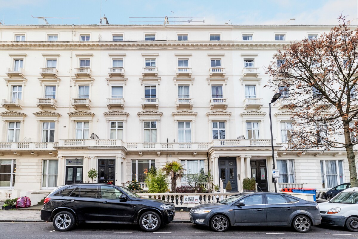 A historic grade two listed building is depicted, featuring a classic white facade adorned with ornate balconies and windows. Two parked cars can be seen in front, along with well-maintained greenery, contributing to the inviting atmosphere of the street.