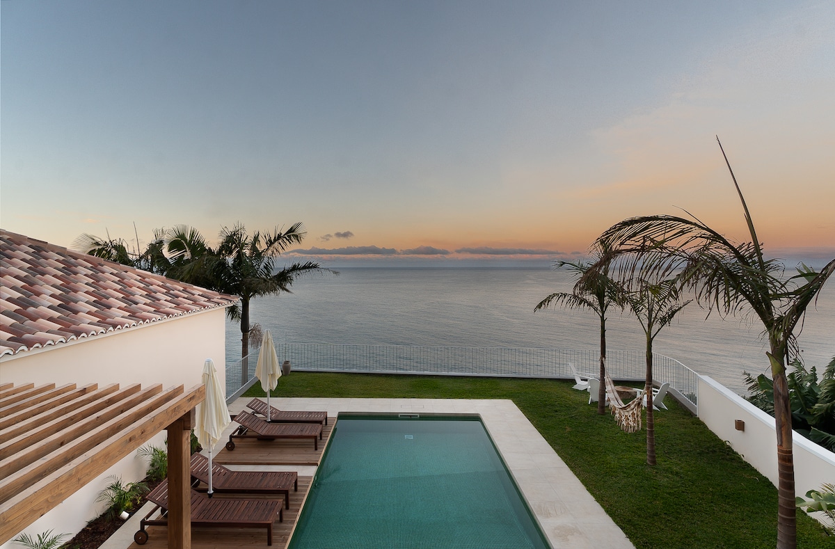 The image showcases an outdoor area featuring a private pool set against a backdrop of the Atlantic Ocean at dusk. Sun loungers are arranged nearby under shade umbrellas, with lush greenery surrounding the space. The horizon displays soft hues of sunset, enhancing the serene atmosphere.