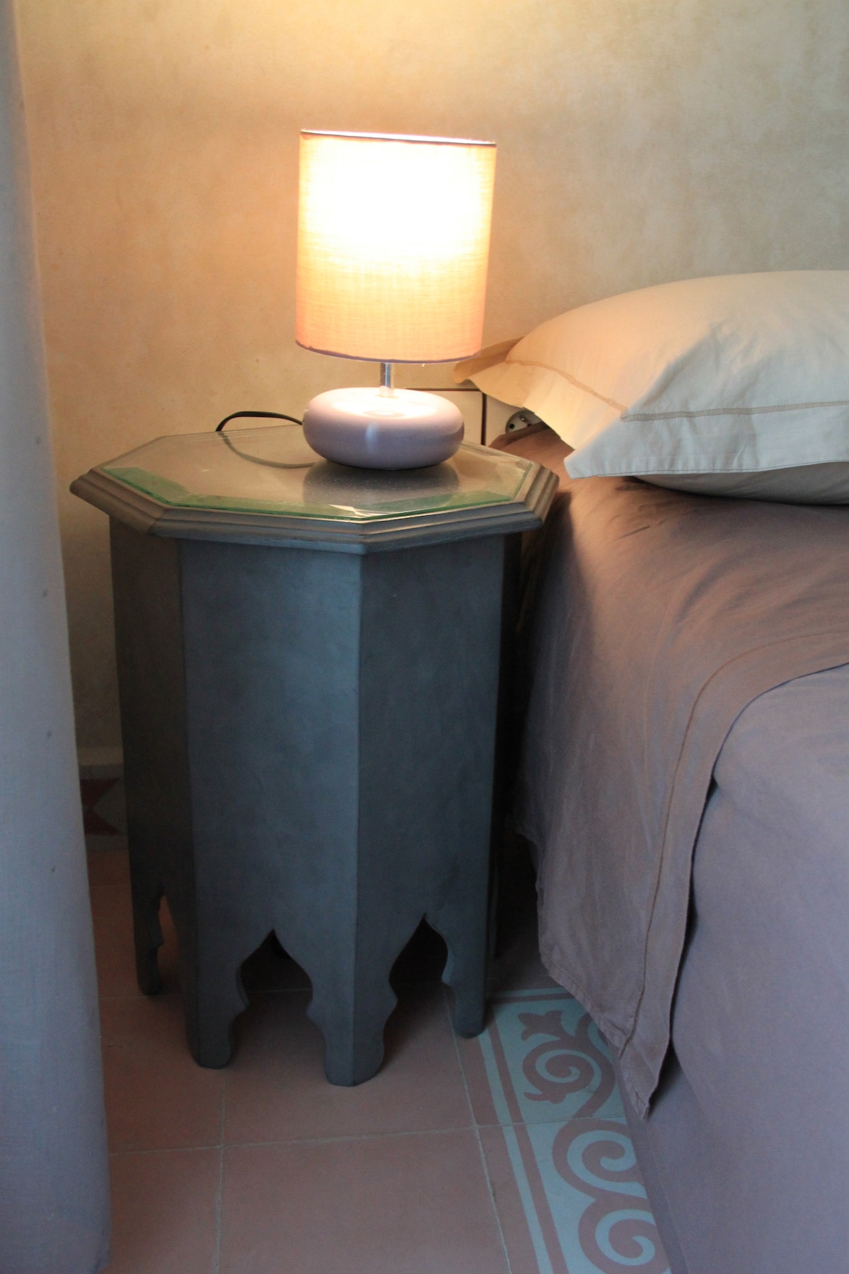 A bedside table with an octagonal shape is positioned next to a spacious bed. A soft lamp emits warm light, illuminating the area. The table's design features traditional elements, with a textured surface and elegant cutouts. Neutral tones are present in the bedding and surrounding walls.