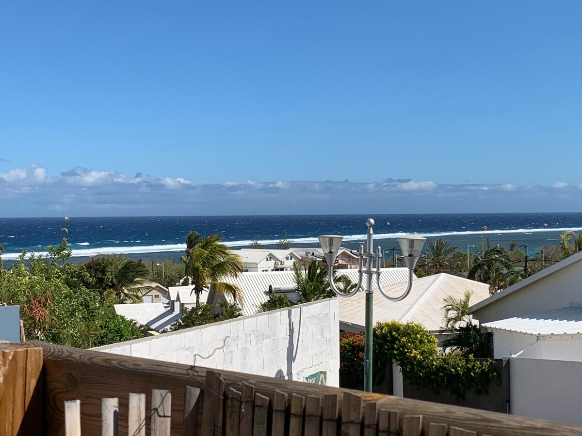 A panoramic view of the ocean is displayed, with vibrant blue waters meeting a clear sky. Palm trees and rooftops from nearby homes can be seen in the foreground, creating a serene coastal atmosphere.