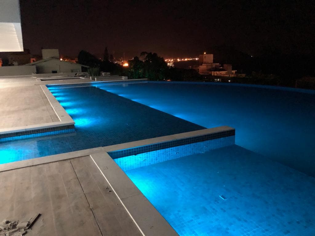 An illuminated infinity pool is showcased at night, with gentle blue lights enhancing the water's surface. The pool area is surrounded by a spacious deck, offering a view of the distant city lights under the night sky.