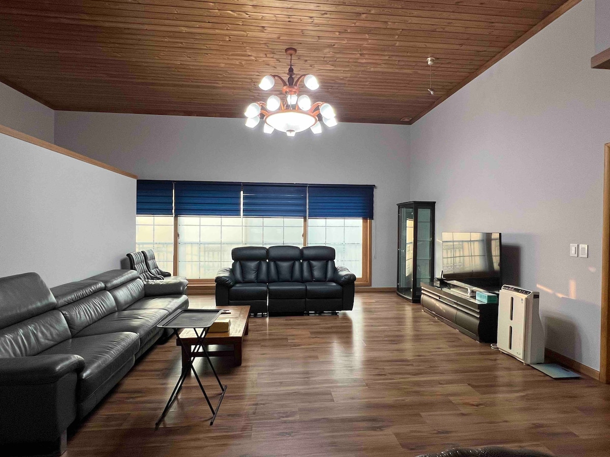 A spacious living room features a high wooden ceiling and large windows adorned with blue blinds. Two black leather sofas provide comfortable seating, complemented by a coffee table. A television is positioned beside a glass cabinet, creating an open and inviting atmosphere.