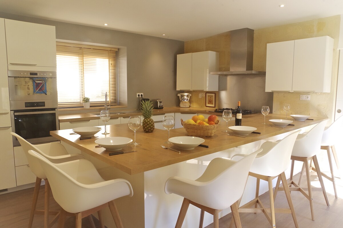 The kitchen features a spacious island with bar seating for eight, complemented by white chairs. Natural light filters through the window, illuminating the modern cabinetry and appliances, including an oven and microwave. A fruit basket and wine bottle add a touch of hospitality.