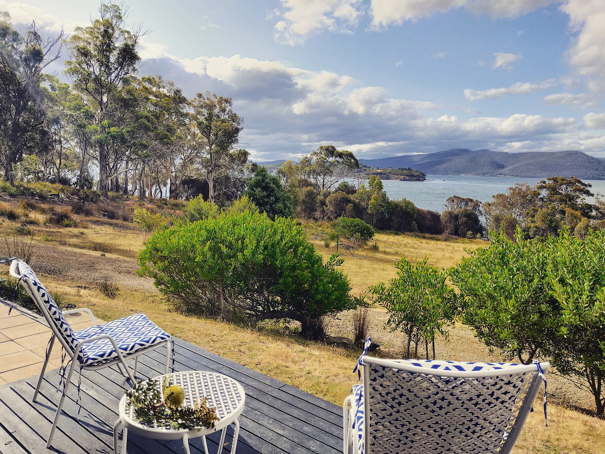 A spacious outdoor deck features two stylish chairs and a small table, overlooking a scenic landscape. Lush greenery frames the view of the waterfront, with rolling hills and distant mountains enhancing the serene atmosphere. Clouds drift across a bright sky, reflecting the natural beauty.