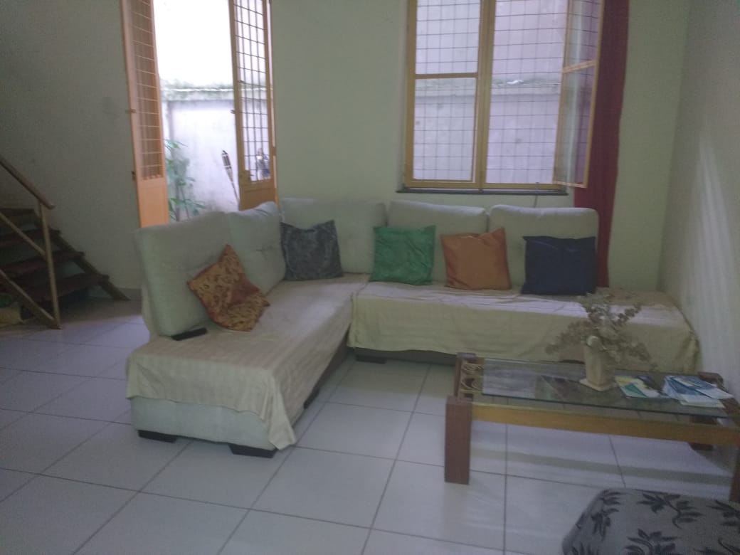 A spacious living area features a large sectional sofa adorned with assorted cushions in shades of green, orange, and gray. A wooden coffee table is positioned in front of the sofa, and natural light enters through two windows with wooden frames, highlighting the tiled floor.