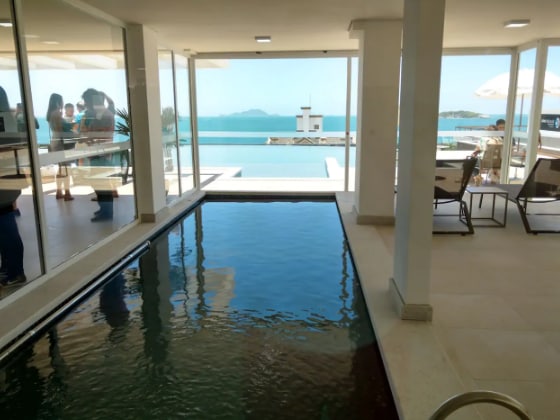 A swimming pool is situated indoors, reflecting natural light from surrounding glass walls. The serene water contrasts with the outdoor view of the sea and deck area furnished with umbrellas and seating. The open space enhances the connection between indoor and outdoor environments.
