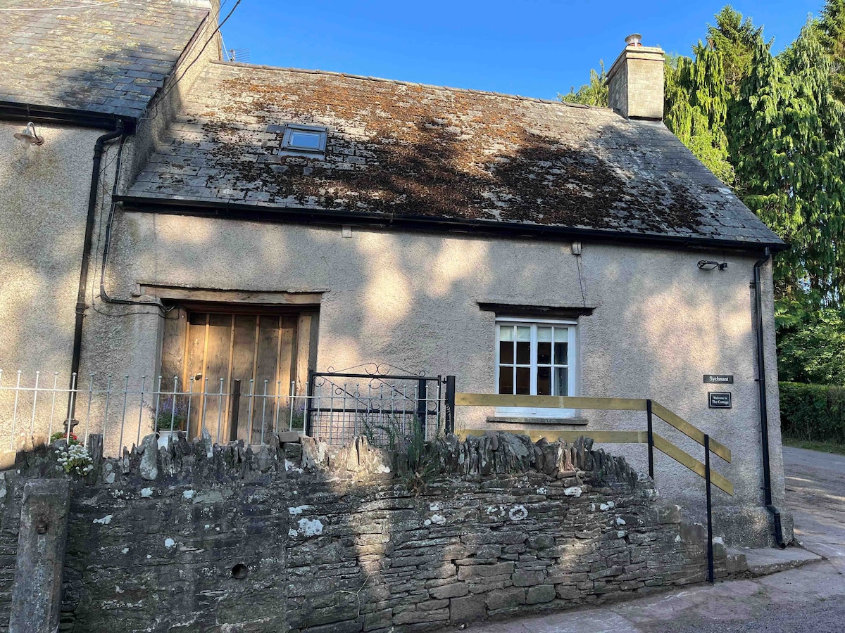 The exterior of The Cottage presents a charming building with a traditional stone façade and a slate roof. A wooden door is centrally located, framed by a quaint fence and natural landscaping. The atmosphere is peaceful, accentuated by surrounding trees and clear blue skies.