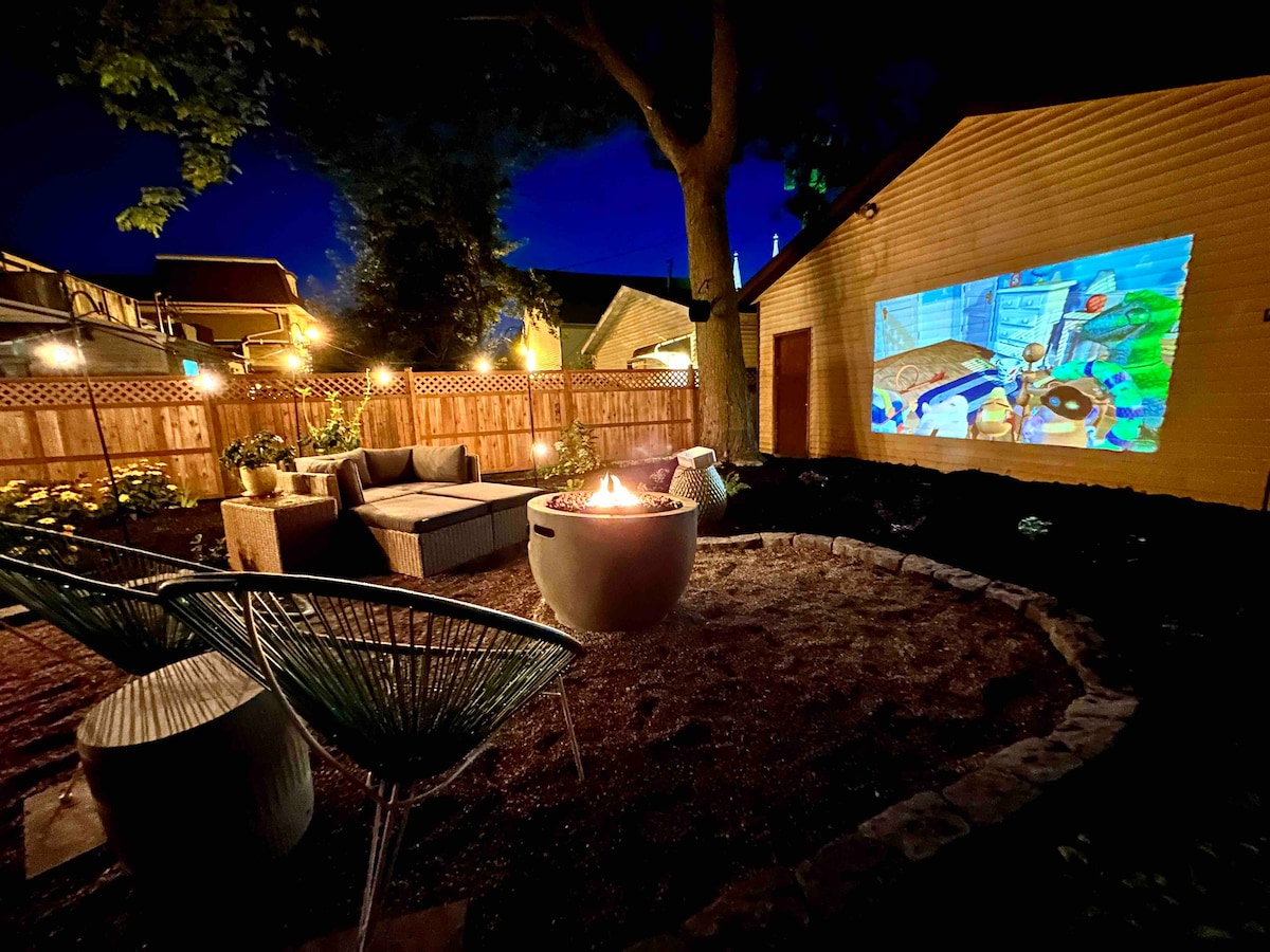 A cozy backyard oasis is illuminated in evening light, featuring comfortable seating around a fire pit. An outdoor movie screen projects a vibrant image onto the adjacent wall, surrounded by lush landscaping and wooden fencing that enhances privacy.