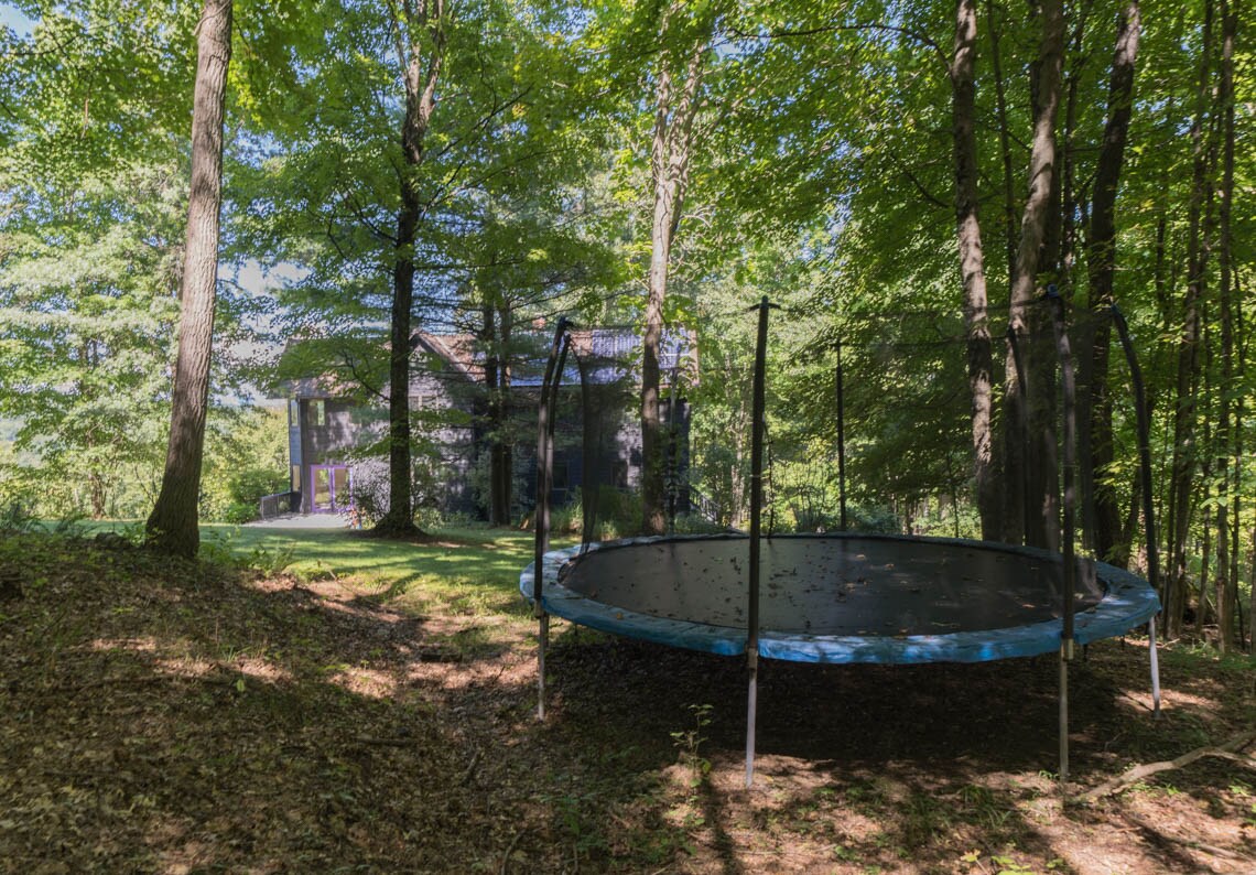 A large trampoline is situated among the trees, surrounded by a natural woodland setting. The contemporary home is visible in the background, partially obscured by lush foliage, offering a serene and private outdoor space.