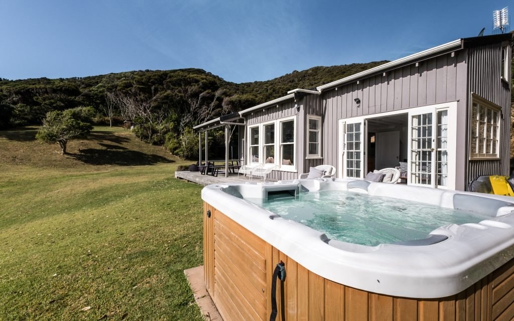 A spa pool is positioned outside a cottage, with the setting sun reflecting off the water. The structure features multiple doors leading to outdoor seating options. Lush green hills rise in the background, showcasing the natural surroundings of the property.