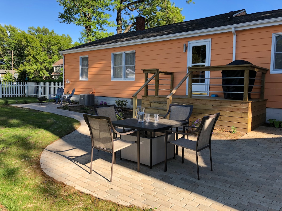 A private backyard features a patio space with a table surrounded by four chairs. The area is paved with stones and includes a raised wooden deck, a grill on the deck, and flower beds adding a touch of greenery. The house is painted in a warm peach color.