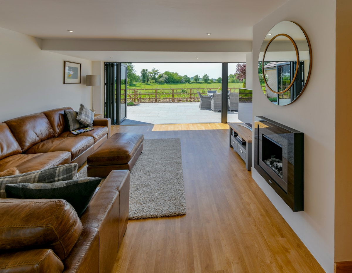 A spacious lounge area features leather sofas arranged around a soft area rug. Large bi-fold doors lead to a patio with views of the green countryside, while a round mirror and a modern fireplace add subtle elegance to the space.