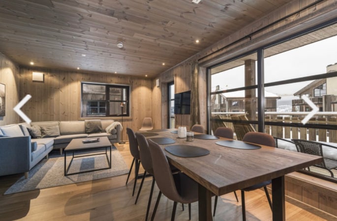 An inviting living space features wooden walls and a cozy seating area with a light-colored sofa. A dining table with chairs offers ample seating. Large glass doors provide views of the outside, enhancing the connection to the outdoors.