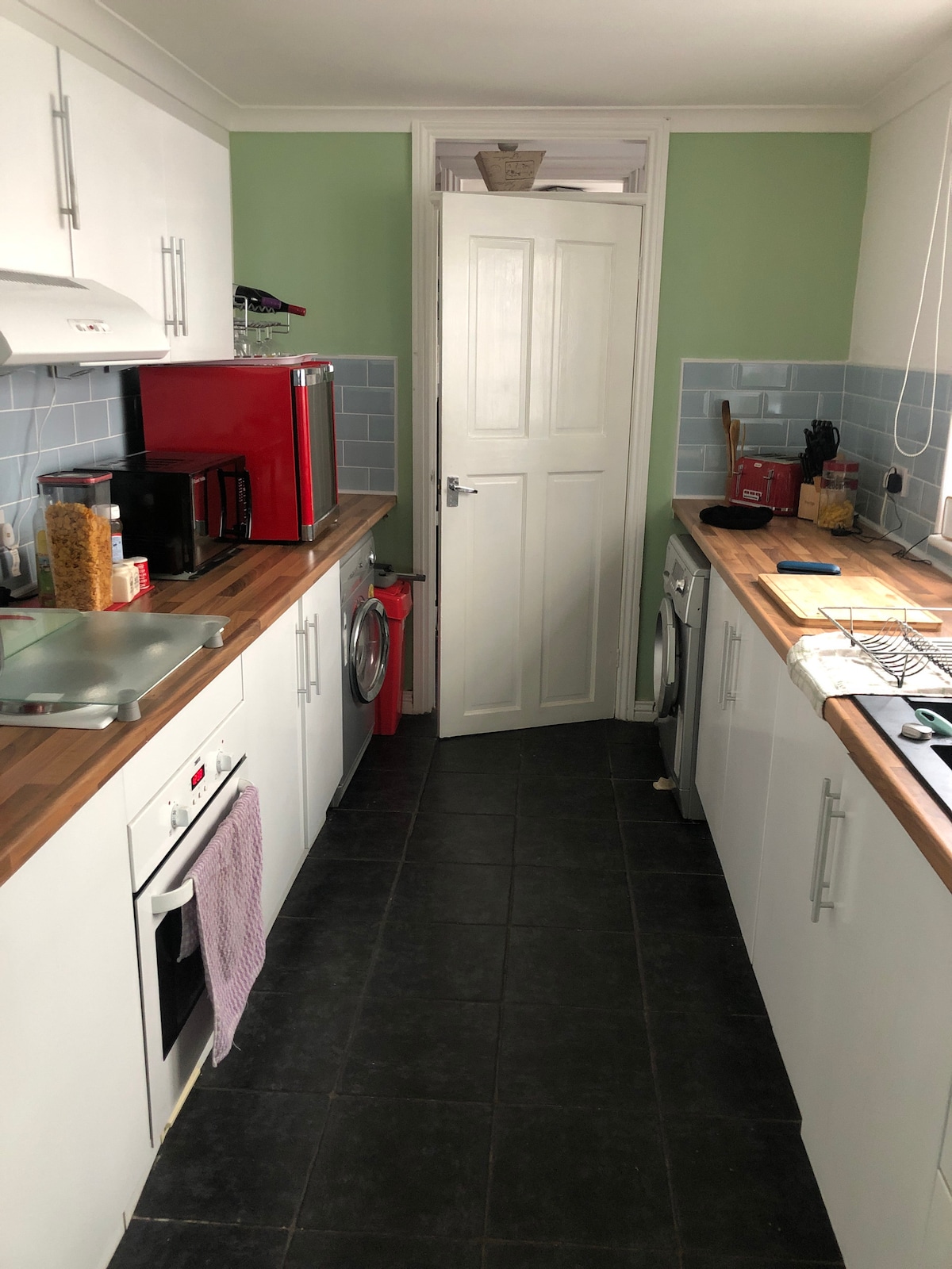 The kitchen features modern appliances, including a red refrigerator and a gas stove. White cabinetry contrasts with a wooden countertop, and blue backsplash tiles provide a subtle color accent. A door leads to another area, while the floor is fitted with dark tiles.