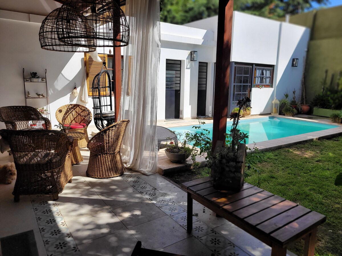 An outdoor seating area features woven chairs and a wooden table, positioned near a tranquil pool surrounded by greenery. Natural light filters through sheer curtains, adding a serene ambiance to the space, which connects with the interior of the house.