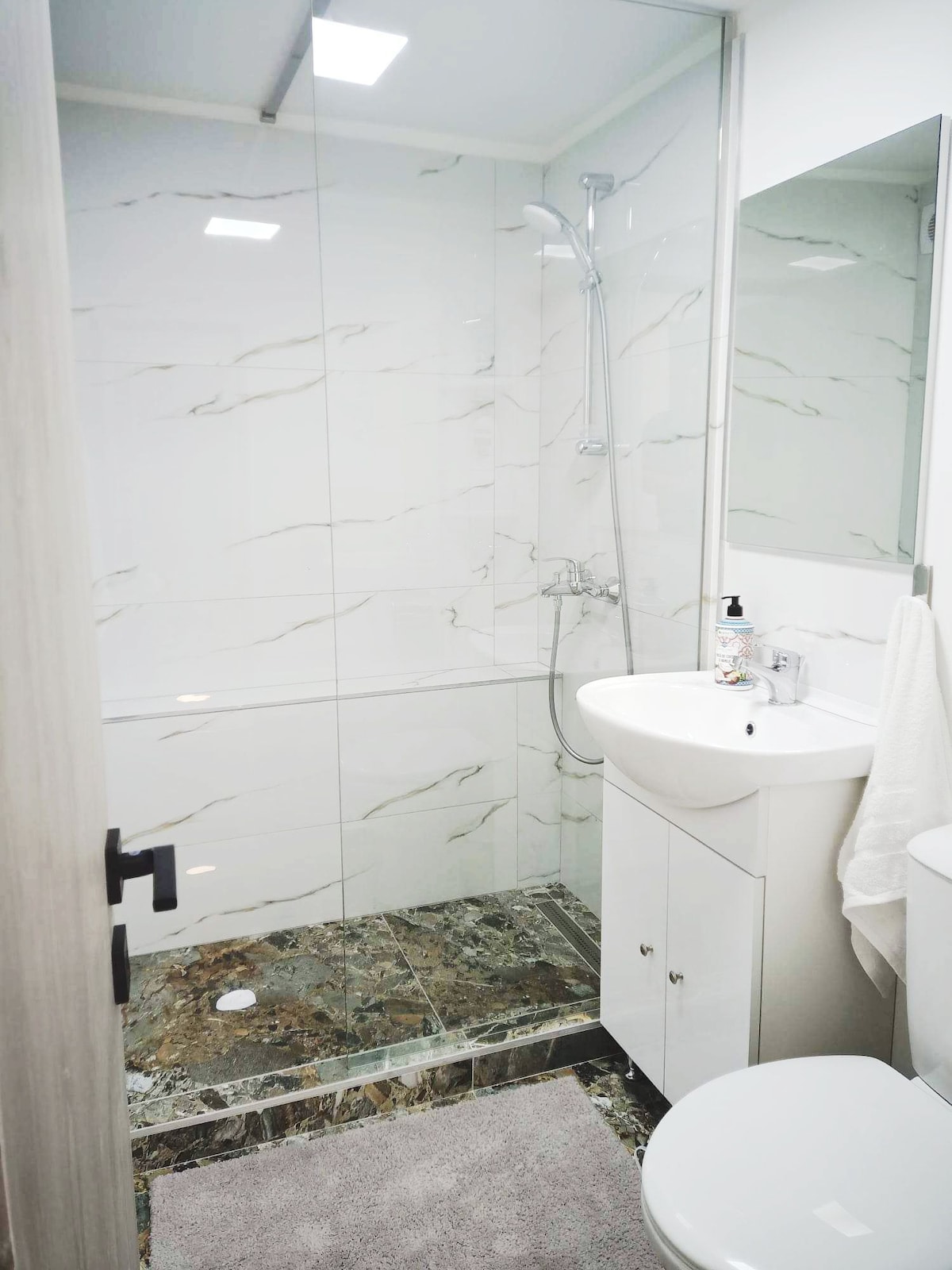 The private bathroom is equipped with a shower and features modern fixtures. A sink with storage underneath, and a toilet are visible. Light marble tiles cover the walls, while the floor showcases a darker patterned design. Complimentary toiletries are neatly arranged on the sink.