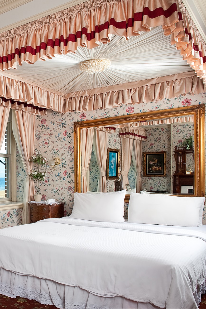 A well-appointed room displays a spacious bed with crisp white linens positioned against floral wallpaper. Elegant draping adorns the ceiling, and a golden-framed mirror reflects the serene ambiance. A glimpse of the ocean view can be seen through the window, enhancing the relaxing environment.