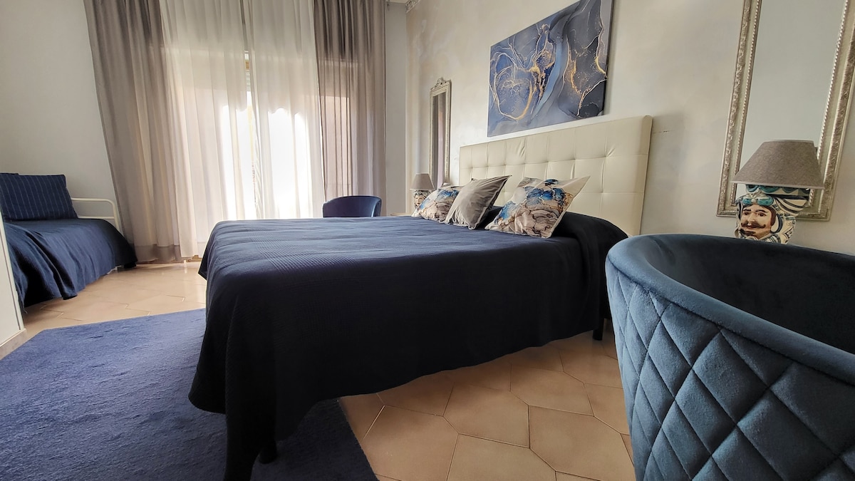 A modern bedroom features a spacious bed adorned with decorative pillows in a color palette reflecting the sea. Natural light filters through sheer curtains, illuminating the room. A cozy seating area is visible, along with elegant mirrors enhancing the room's dimensions.