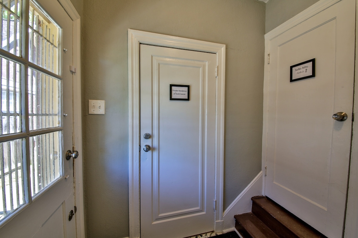 The entryway features a solid door with a notification sign mounted on it, alongside a staircase leading to the upper floor. Natural light enters through a glass-paneled door, illuminating the space, while light-colored walls provide a simple, clean backdrop.