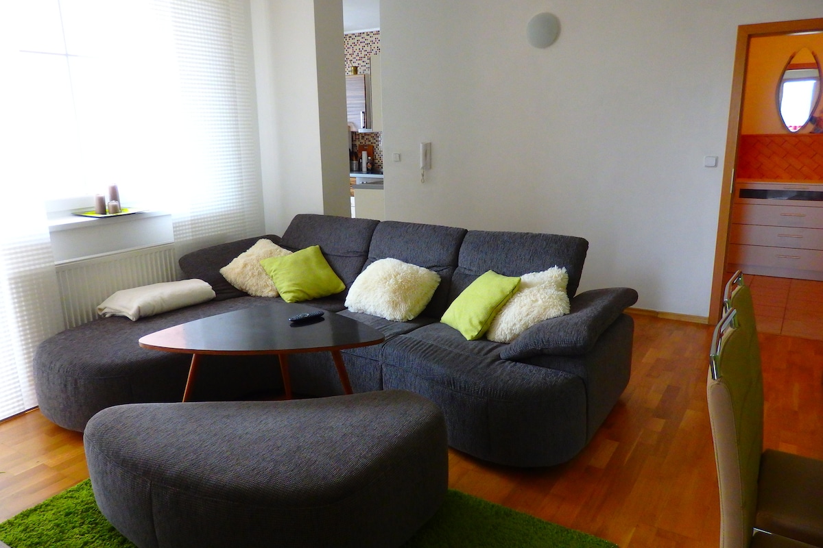 A spacious living area features a modern, curved sofa adorned with decorative cushions. A low coffee table rests in front, accompanied by a grassy area rug. Natural light fills the space through large windows, while a dining area with seating is visible in the corner.