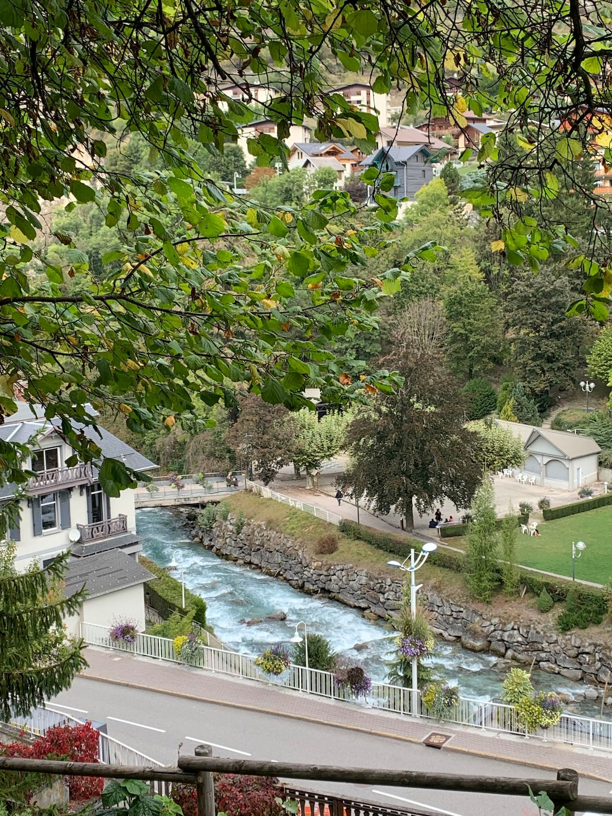 A view of a flowing river bordered by greenery and walking paths. Nearby, buildings are nestled among trees, while the surrounding landscape showcases manicured lawns and natural foliage, providing a serene atmosphere.
