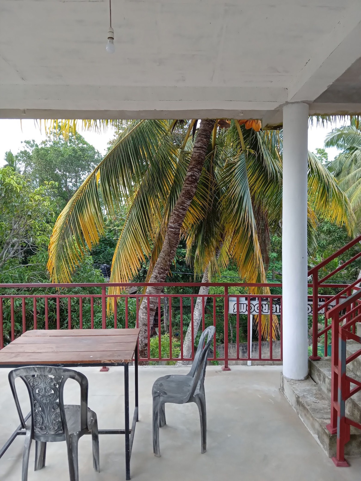 A balcony area is presented with a simple table and two chairs, offering a relaxed space for enjoying the outdoor view. Lush green trees and palm fronds create a natural backdrop, enhancing the connection to the surrounding environment.