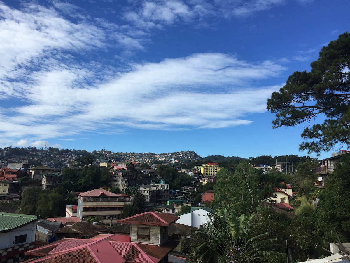 A view of a residential neighborhood under a clear blue sky with wispy clouds. Various houses with red roofs and green exteriors are visible, set against a backdrop of hills and trees, indicating a serene and inviting atmosphere.