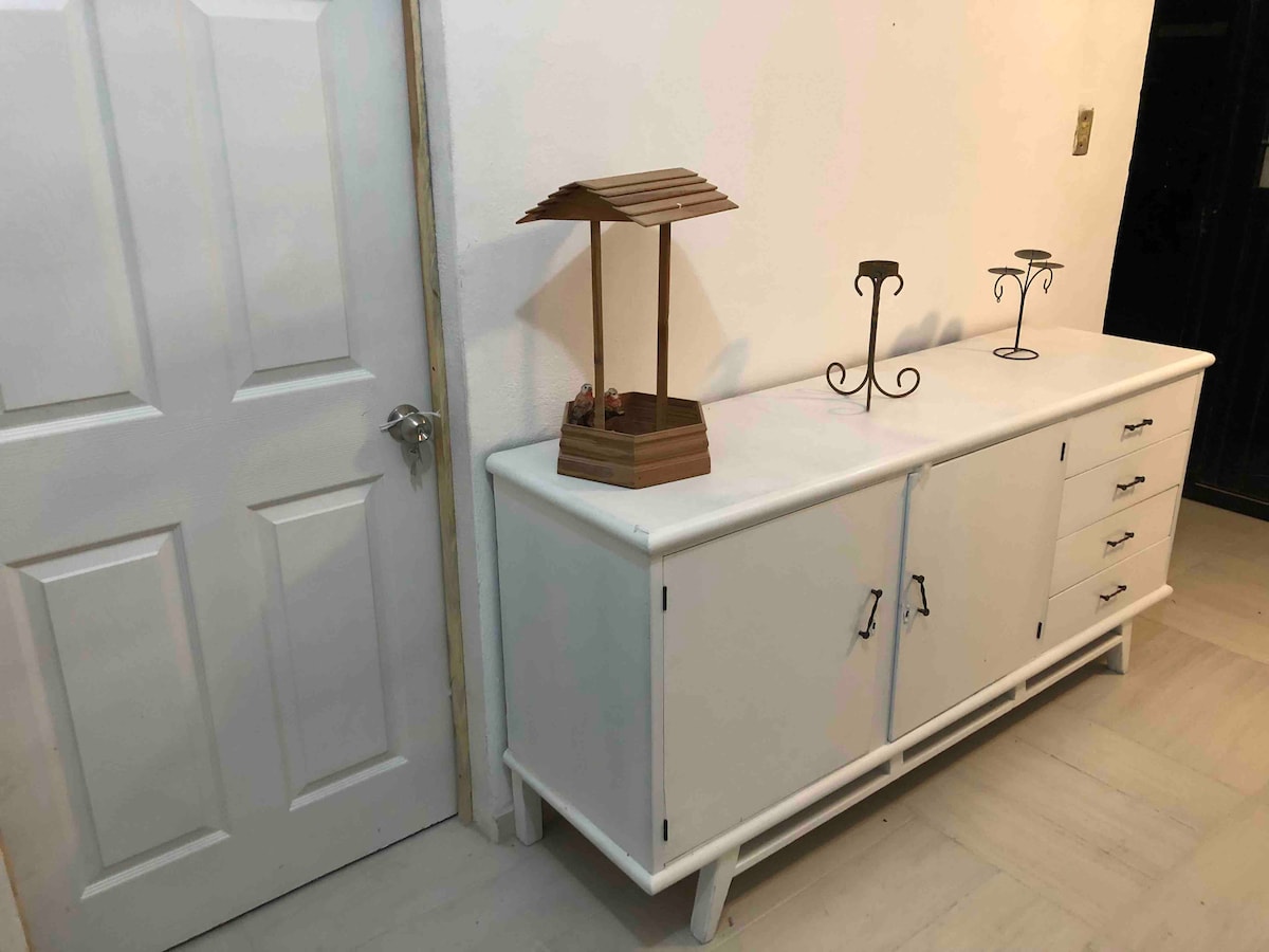 A white sideboard is positioned against a wall, featuring doors and drawers for storage. Two decorative candle holders stand alongside a wooden lantern on top, contributing to the room's functional decor. A closed door is visible nearby.