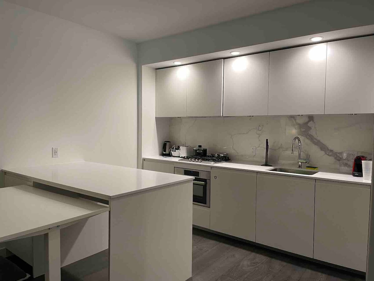 A sleek kitchen is designed with modern cabinetry and a light countertop. Essential appliances include a gas stove, and a refrigerator, and a small dining area with counter seating is visible. Ambient lighting enhances the clean lines and minimalistic style of the space.