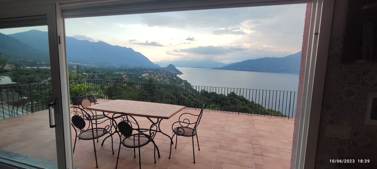 A spacious outdoor terrace is visible, featuring a rectangular table surrounded by six metal chairs. The scene offers a panoramic view of the lake and mountains under a cloudy sky, creating a serene backdrop for relaxation.