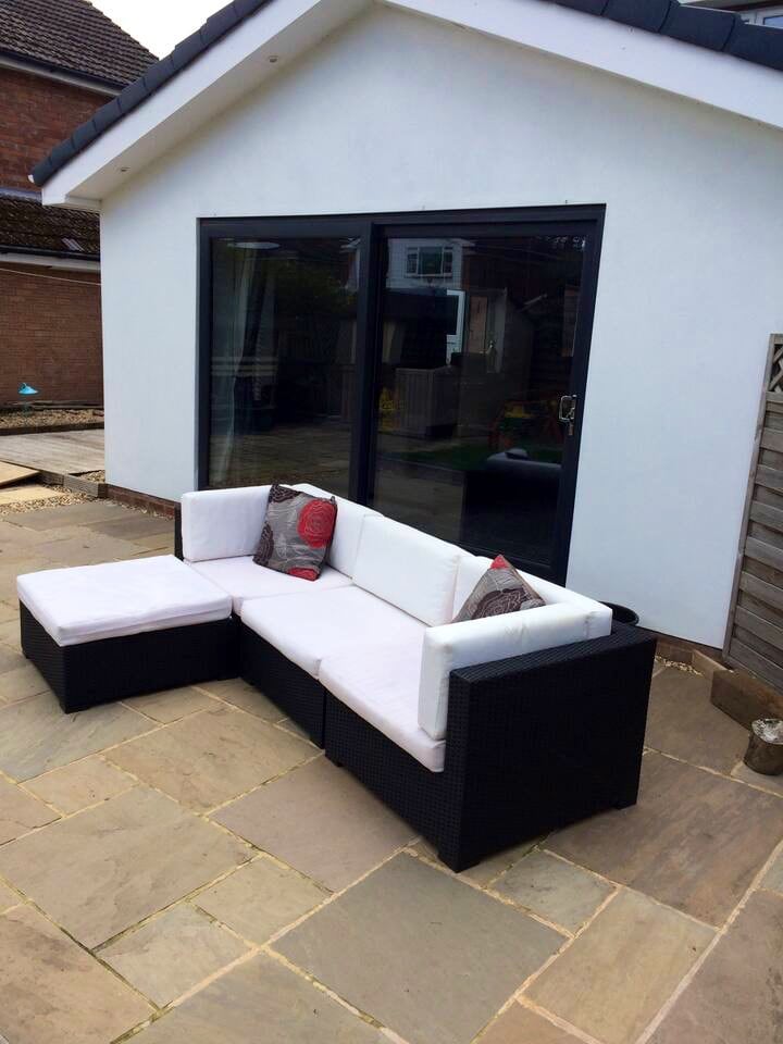 An outdoor lounge area is featured, showcasing a sectional sofa with white cushions and decorative pillows. The sofa is positioned on a stone patio, with large glass doors in the background providing access to the interior space.