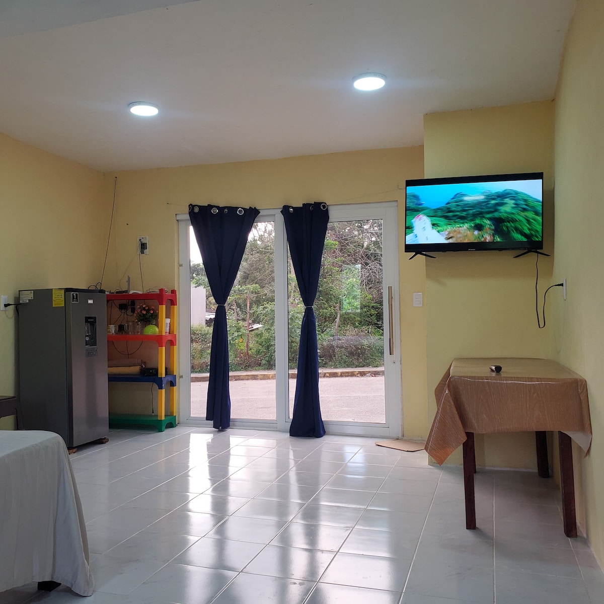 An inviting living space features a large window with blue curtains, allowing natural light to filter in. A flat-screen television is mounted on the wall, while a wooden table is positioned nearby. A refrigerator is located in the corner, alongside a colorful shelving unit.
