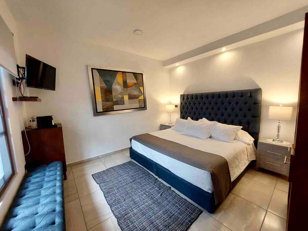 A well-appointed room is displayed, featuring a spacious bed with an upholstered headboard. Modern bedside lamps provide soft lighting beside the bed. A colorful artwork hangs above the furniture, and a flat-screen television is mounted on the wall, creating a comfortable ambiance.