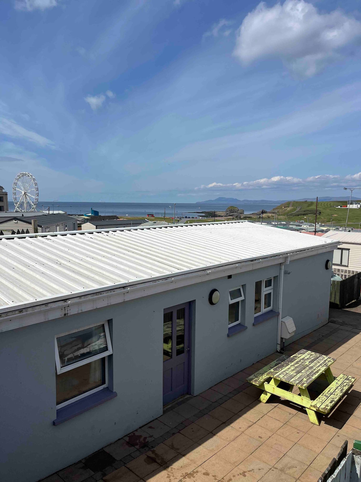 A view from above showcases a light-colored building with multiple windows and a purple door. The patio area is furnished with a picnic table. In the background, the ocean and a Ferris wheel are visible against a cloudy sky.