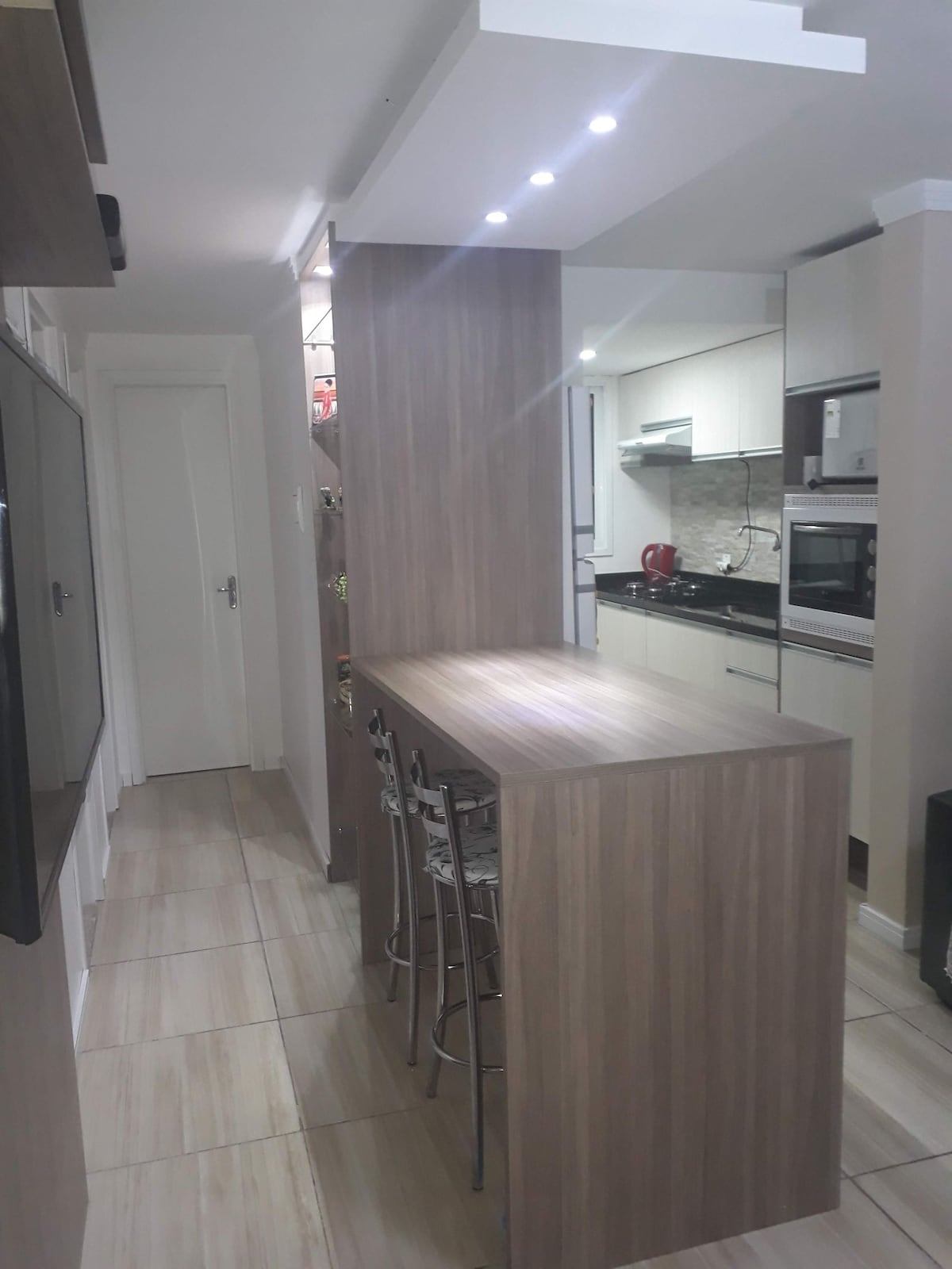 A modern kitchen features sleek cabinetry and a long, wooden island with four barstools. Open shelving displays delicate glassware, and appliances are subtly integrated into the design. Bright lighting highlights the clean lines of the space, creating a functional and inviting area.