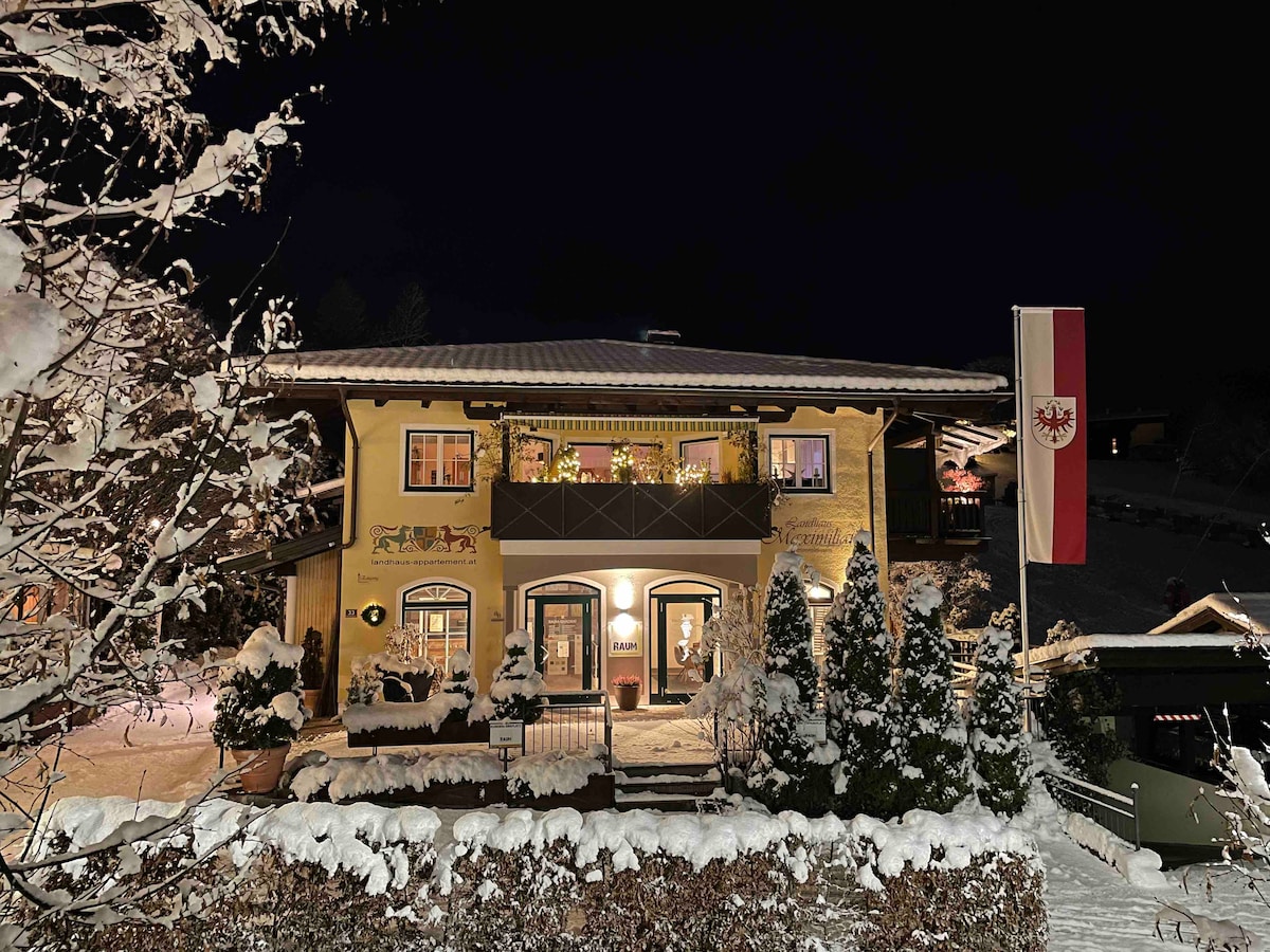 The charming exterior of the vacation home is captured at night, adorned with a fresh layer of snow. Warm light spills from the windows, and a flag waves gently in the winter breeze. Lush greenery and decorative elements frame the inviting entrance.