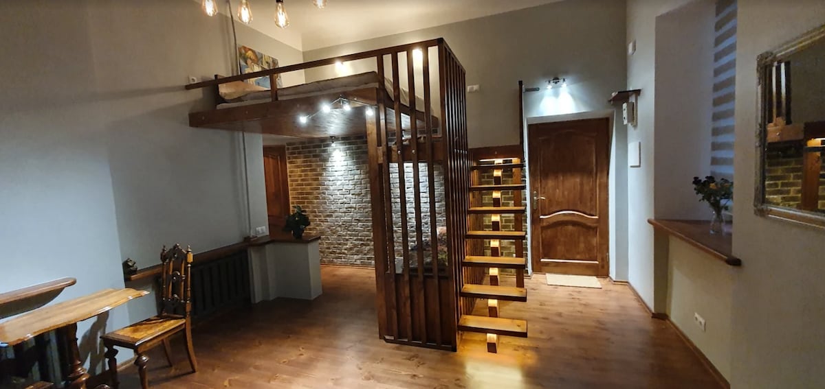 An open living space features a wooden staircase leading to a second-level area. Natural light is provided by recessed ceiling fixtures and a wall of illuminated bricks. A wooden dining table is visible on one side, with a charming wooden chair positioned beside it.