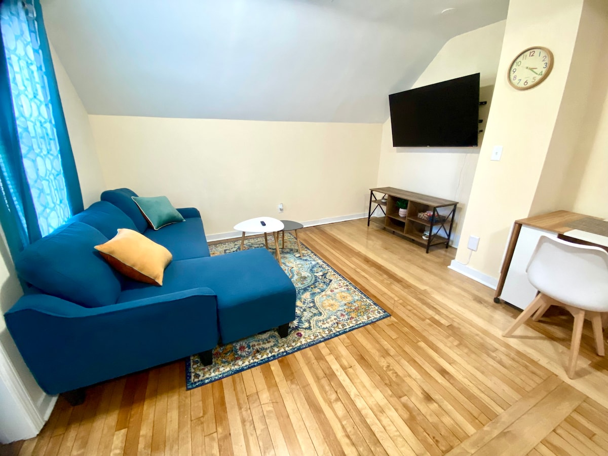 A living area is presented with a blue sectional sofa adorned with colorful cushions. A round coffee table is situated in front. A large wall-mounted TV is visible, complemented by a wooden side table and a clock on the wall. Natural light enters through a window with patterned curtains.