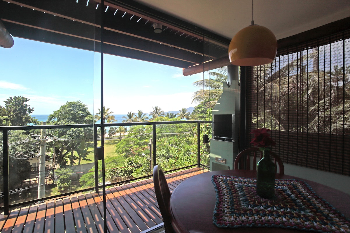 A dining area features a round table with a central vase holding a flower. Large glass doors provide an unobstructed view of the ocean and surrounding greenery, creating a bright and airy atmosphere. Natural light floods the space, enhanced by the presence of a pendant light overhead.