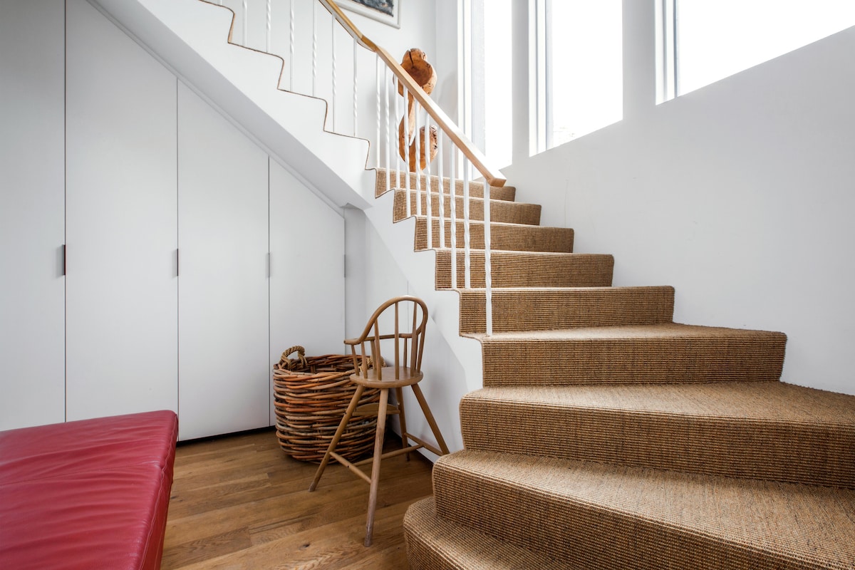 A flight of carpeted stairs leads up with white wooden railings alongside. Natural light filters through large windows, highlighting a wooden chair positioned beside a woven basket. Closets with white doors are visible in the background, providing practical storage options.