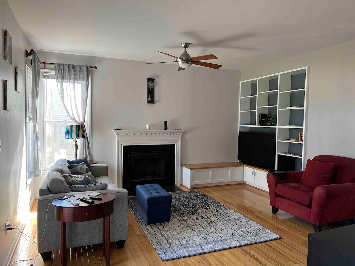 A spacious living area features a comfortable gray sofa and a red armchair, providing ample seating. A neutral color palette is complemented by a patterned area rug on hardwood flooring. Light streams in through large windows, highlighting the cozy ambiance.