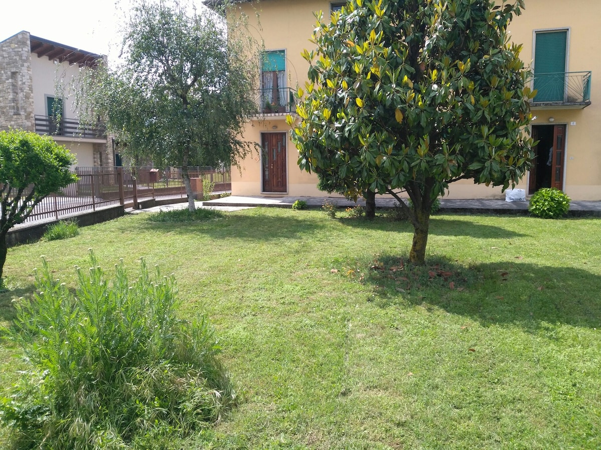 A spacious garden is visible, featuring neatly trimmed grass and several trees, including a large magnolia. The exterior of a two-story building can be seen in the background, with multiple entryways and a balcony. The area appears bright and inviting.
