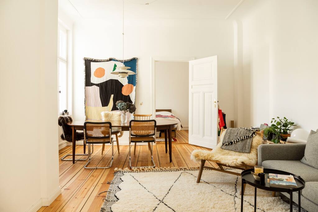 A bright, airy space showcases a dining area with a wooden table and four chairs. A colorful textile artwork adorns the wall. A cozy living area features a gray sofa and a small side table, positioned on a patterned rug across the polished wooden floor.