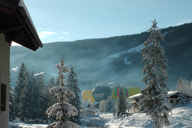 Snow-covered trees frame a winter landscape, highlighting the distant view of colorful hot air balloons rising against a backdrop of misty mountains. Soft sunlight filters through the scene, creating a serene winter atmosphere in the beautifully snowy surroundings.